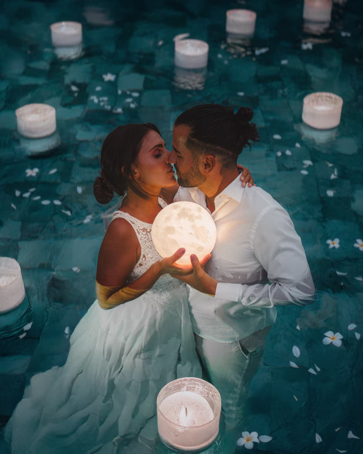 Los novios besándose en una piscina de noche sosteniendo una luna encendida entre velas flotantes