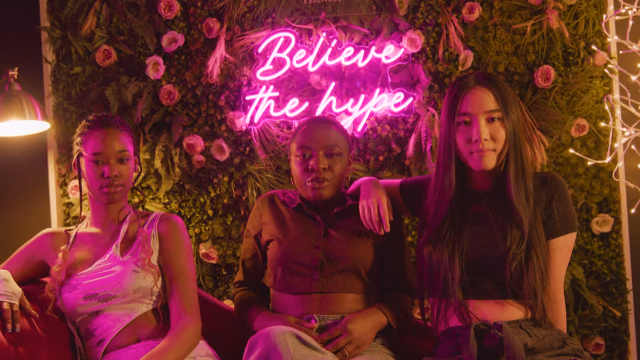 Three women seated on a sofa beneath a pink 'Believe the hype' neon sign on a flower wall at an Afroroots club night