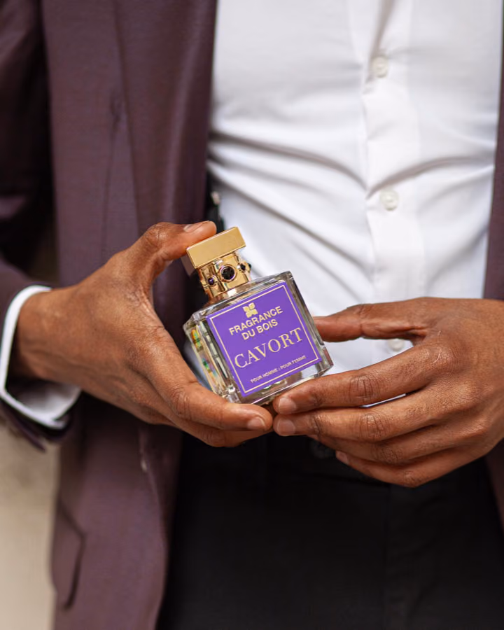 Model in a plum suit holding a Fragrance Du Bois Cavort perfume bottle with purple label and gold cap