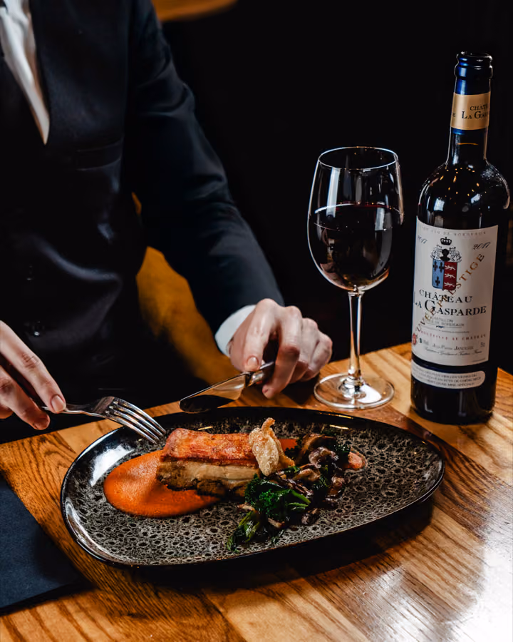 Diner in a dark suit cutting into crispy pork belly with romesco sauce beside a bottle of Bordeaux at West End Brasserie, Edinburgh